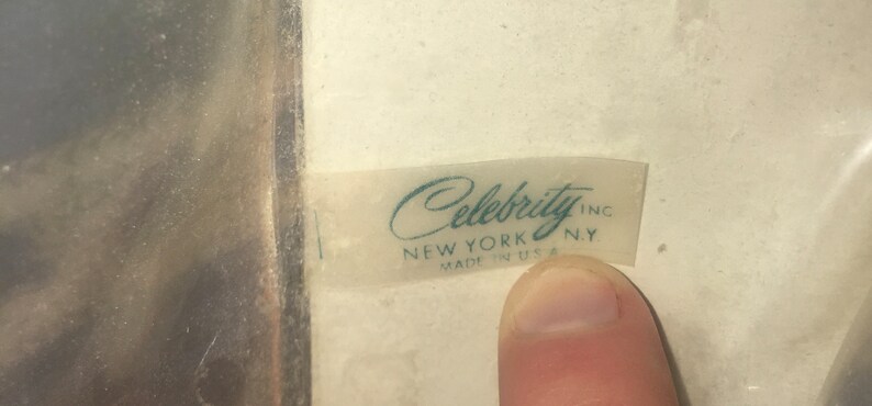 Op de afbeelding: Een wit label met de tekst "Celebrity Inc. New York, NY Made in USA" in turquoise letters.