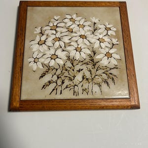 May include: A square ceramic tile featuring a hand-painted illustration of a cluster of white daisies with yellow centers and dark brown stems and leaves. The tile is encased in a simple, medium-toned wooden frame with a visible wood grain texture.