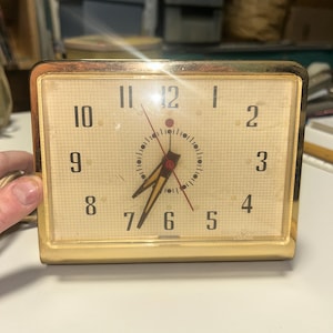 May include: A vintage rectangular alarm clock with a gold-colored frame. The clock face is beige with black numerals and hands. The clock shows the time as 6:58. The clock has a power cord.