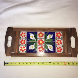 May include: A wooden serving tray with a tile inlay featuring a repeating pattern of red flowers, white calla lilies, and green leaves on a blue background. The tray has two wooden handles.
