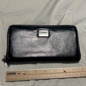 May include: A black rectangular zip-around wallet with a textured finish and a silver-toned metal plaque featuring the text ESPRIT SAN FRANCISCO. A wooden ruler is placed along the bottom edge, showing the wallet is approximately 8 inches long.