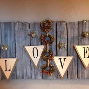 May include: A rustic wooden sign with the word "LOVE" spelled out in white triangular pennants. The pennants are hung with twine and have black lettering. The background is a weathered grey wood plank.