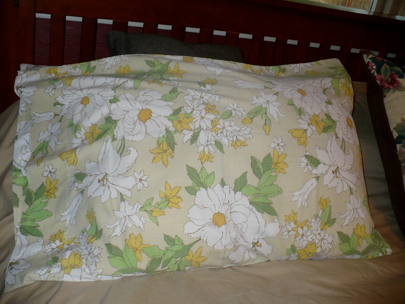 Puede incluir: Una almohada con un patr&oacute;n floral que presenta flores blancas con centros amarillos sobre un fondo beige claro. Las flores est&aacute;n rodeadas de hojas verdes.