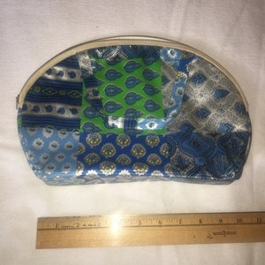 Op de afbeelding: Een witte, halfronde, met een rits te sluiten tas met een patchwork-ontwerp. De patchwork is gemaakt van blauwe, groene en witte stof met verschillende patronen. De tas is ongeveer 18 cm breed.