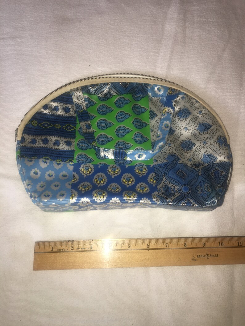 Op de afbeelding: Een witte, halfronde, met een rits te sluiten tas met een patchwork-ontwerp. De patchwork is gemaakt van blauwe, groene en witte stof met verschillende patronen. De tas is ongeveer 18 cm breed.
