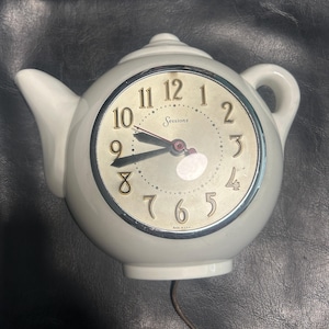 May include: A white ceramic teapot-shaped wall clock with a round face featuring gold-coloured numbers from 1 to 12. The clock face displays the brand name Sessions in script, black hour and minute hands, a red second hand, and Made in U.S.A. text.