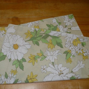 May include: A beige fabric with a floral pattern featuring white flowers with yellow centers and green leaves. The flowers are arranged in a repeating pattern.