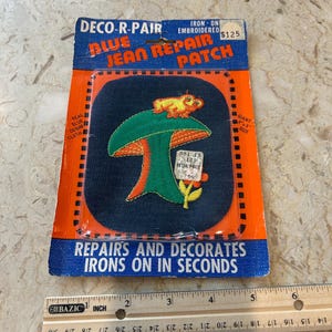 May include: Embroidered blue jean repair patch with a mushroom design and a frog on top. The patch is on a blue and orange card that reads "DECO-R-PAIR BLUE JEAN REPAIR PATCH". The patch measures approximately 4 inches by 5 inches.