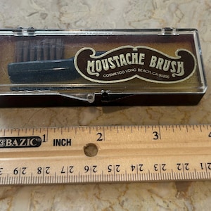 May include: A vintage mustache brush in a clear plastic case. The case is brown with a hinged lid. The brush is black with dark bristles. The label reads "MOUSTACHE BRUSH" and "COSMETCO LONG BEACH, CA 90806". A ruler is next to the case.