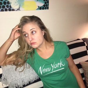 May include: A woman wearing a green t-shirt with the words "New York" written in white on the front. She is sitting on a striped couch with a gray fringed pillow.