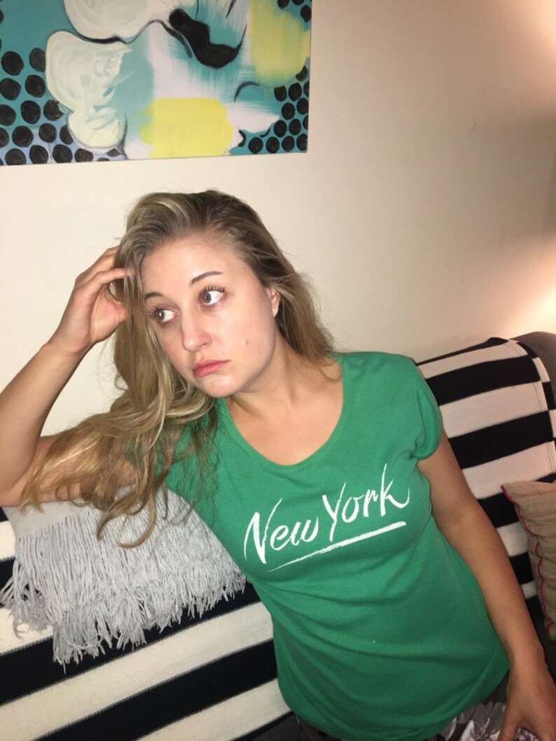 May include: A woman wearing a green t-shirt with the words "New York" written in white on the front. She is sitting on a striped couch with a gray fringed pillow.