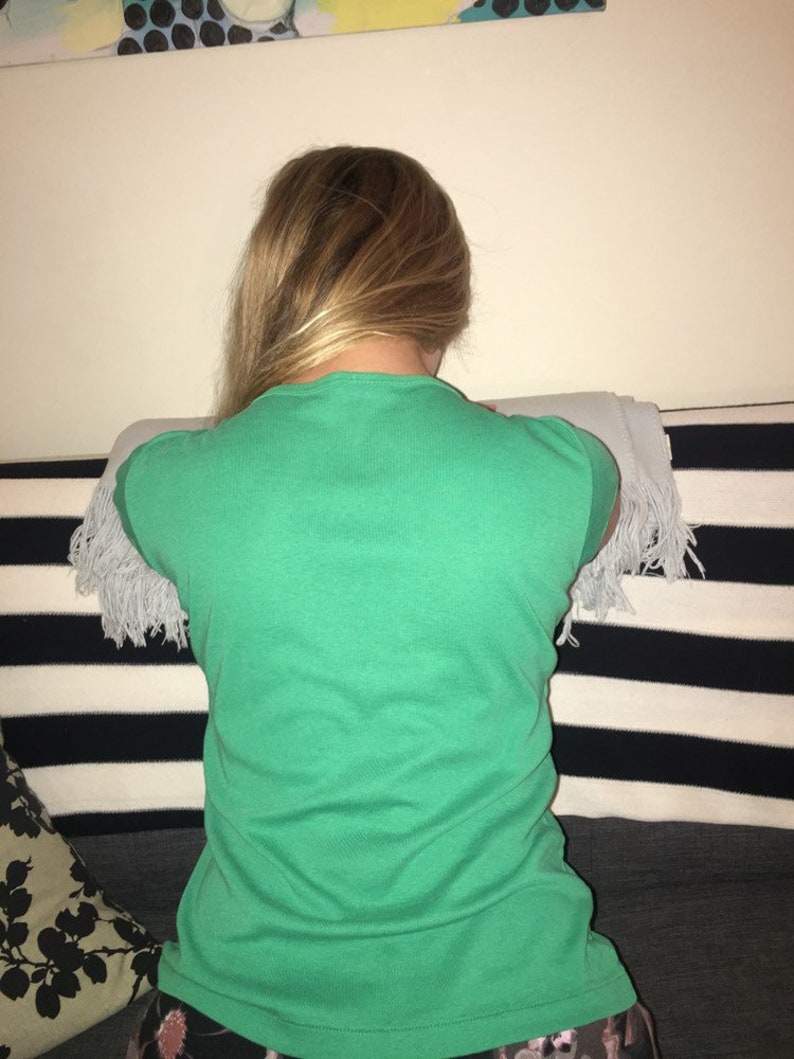 May include: A woman wearing a green t-shirt with a white fringe trim on the sleeves. She is sitting on a striped couch with a black and white pattern.