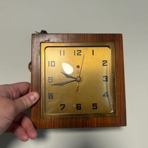 May include: A square wall clock with a dark wood frame and a gold-toned metal face. The clock features black numerals, black hour and minute hands, and a thin gold second hand. A ruler at the bottom shows the clock is approximately 6 inches wide.