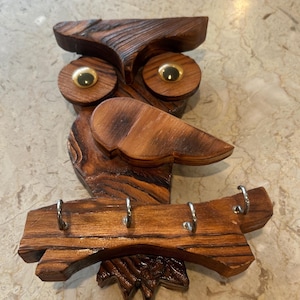 May include: Wooden owl-shaped key holder with a natural wood grain finish. The owl has large, round eyes with gold-colored centers and a prominent beak. Four silver key hooks are attached to a wooden branch below.