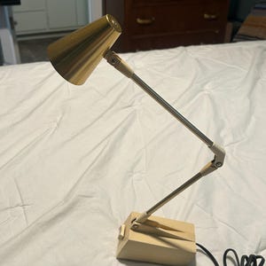 May include: A desk lamp featuring a metallic gold conical shade attached to a two-section adjustable arm. The base and arm joints are finished in a matte cream color. A black power cord extends from the rectangular base, which includes a side switch.