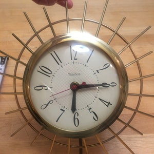 Peut inclure: Une horloge dorée avec un cadran blanc et des aiguilles noires. L'horloge a un design unique avec un cadre circulaire et des rayons qui rayonnent. Le cadran de l'horloge porte l'inscription "United".