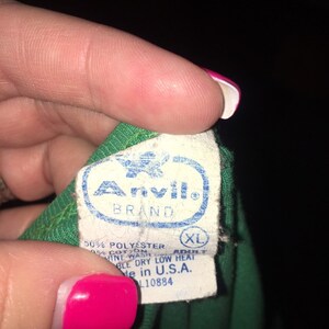 May include: A white tag with black text that reads "Anvil Brand" with a green bird logo. The tag also says "50% Polyester 50% Cotton", "Adult", "XL", "Machine Wash Low Heat", "Tumble Dry Low Heat", "Made in U.S.A.", and "L10884".