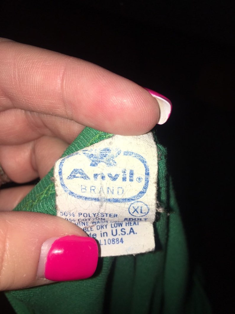 May include: A white tag with black text that reads "Anvil Brand" with a green bird logo. The tag also says "50% Polyester 50% Cotton", "Adult", "XL", "Machine Wash Low Heat", "Tumble Dry Low Heat", "Made in U.S.A.", and "L10884".