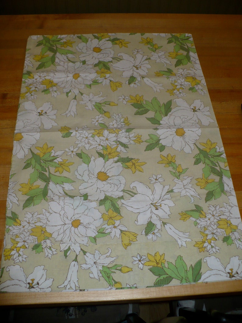 Puede incluir: Una tela beige con un patr&oacute;n floral que presenta flores blancas con centros amarillos y hojas verdes. Las flores est&aacute;n dispuestas en un patr&oacute;n repetitivo.