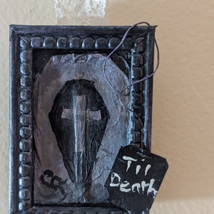 May include: A black framed picture with a silver cross on a gray background. A black tag with white text reads "Til Death".