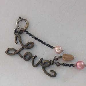 May include: A silver charm with the word "Love" written in cursive, with a chain and pink beads attached.