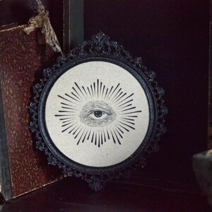 May include: A circular wall hanging with a black frame, white background and a black and white illustration of an eye with radiating lines.