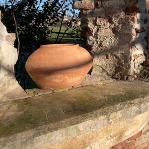 May include: A large, round terracotta pot with a wide body and a narrow neck. The unglazed clay pot sits on a weathered stone ledge, with a backdrop of a crumbling brick wall and foliage. The pot's warm, earthy tones contrast with the cool blue sky.