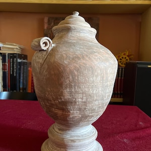Può includere: Un'urna in terracotta con una finitura usurata color marrone chiaro e bianco. Il vaso presenta un corpo arrotondato, una base a piedistallo, un coperchio con un piccolo pomello e un unico manico decorativo a voluta sul lato superiore.
