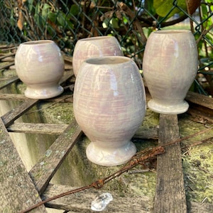 Può includere: Quattro vasi in ceramica fatti a mano con finitura perlata. I vasi sono rosa chiaro e bianchi, con un corpo arrotondato e una piccola base circolare. I vasi sono disposti su una superficie di legno stagionato, con uno sfondo verde.