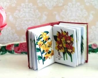 May include: A miniature red book with colorful flower illustrations is open on a white table with two drawers. A small glass vase with pink flowers sits on the table. A gold coin is on the table to the right of the book.