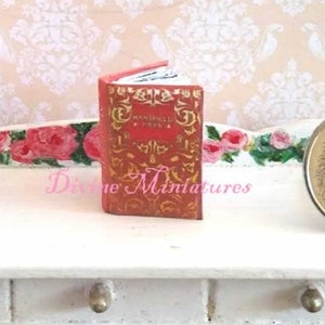 May include: A miniature red book with gold lettering titled "The Wonderful Wizard of Oz" on a white table with two drawers. A gold coin with a design of a bird is on the table to the right of the book.