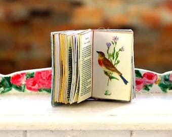 Puede incluir: Un libro en miniatura abierto con una ilustración de un pájaro, apoyado sobre una mesa blanca con detalles florales. Las páginas del libro son visibles, y la mesa tiene dos cajones con pomos de latón. El texto "Divine Miniatures" está en la parte superior.