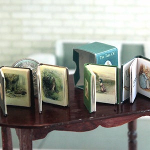 May include: Four miniature books with colourful covers and illustrations. The books are titled "The Tale Of" and feature a rabbit, a squirrel, and a bird. The books are arranged on a wooden table.