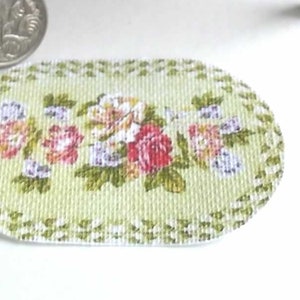 May include: A miniature oval-shaped rug with a floral design. The rug is green with pink, white, and yellow flowers. The rug is about 2 inches long and 1 inch wide.