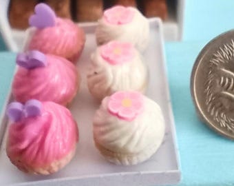 Peut inclure: Présentoir de boulangerie miniature avec des cupcakes et des pâtisseries. Des cupcakes roses et blancs avec des décorations de fleurs et de nœuds sont sur un plateau. Une pièce de monnaie est à côté de l'étalage. Des étagères contiennent des pains et des croissants miniatures.