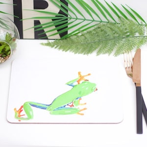 Op de afbeelding: Witte onderzetter met een illustratie van een groene en oranje kikker. De kikker is afgebeeld op een witte achtergrond.