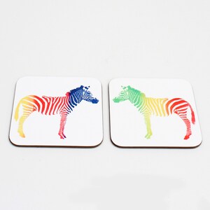 May include: Two square coasters with a white background and a rainbow zebra design. The zebra on the left coaster is blue, red, yellow, and orange. The zebra on the right coaster is green, yellow, orange, and red.