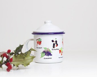 May include: White enamel mug with a blue rim and lid. The mug has a design of different berries with their names printed below: Redcurrant, Elderberry, Blackberry, Wild Strawberry.