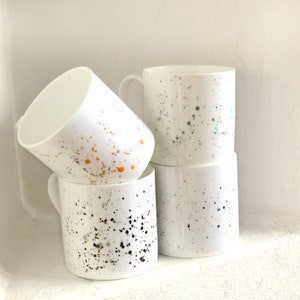 May include: Four white ceramic mugs with different coloured speckles. The mugs are stacked on top of each other. The top mug has green speckles, the second mug has orange speckles, the third mug has black speckles, and the bottom mug has a mix of colours.