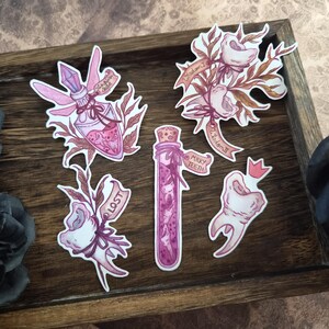 Könnte beinhalten: Eine Sammlung skurriler Aufkleber mit einer Zaubertrankflasche, Zähnen und floralen Elementen. Die Aufkleber sind in Rosa-, Lila- und Brauntönen gehalten und tragen Textaufschriften wie "Milky Teeth" und "Lost". Auf einem Holztablett angeordnet.