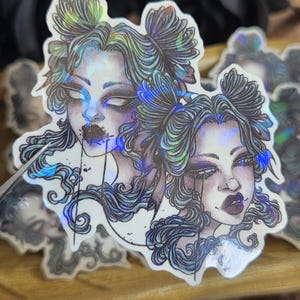 Könnte beinhalten: Holografischer Aufkleber mit einer detaillierten Illustration eines Frauengesichts mit dunklem Make-up und fließendem Haar. Der Aufkleber hat ein reflektierendes, irisierendes Finish mit blauen, grünen und violetten Farbtönen. Das Kunstwerk hat ein gotisches oder Fantasy-Thema.