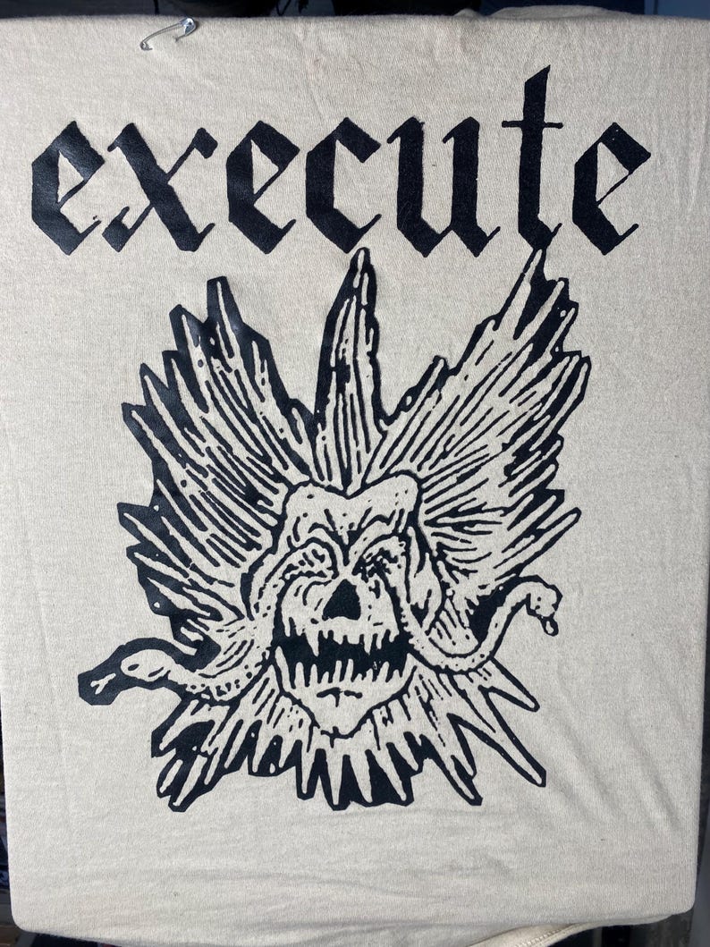 May include: Cream-coloured t-shirt featuring the word "EXECUTE" in a gothic font above a black and white Medusa-style graphic. The graphic shows a skull with snakes and spiky details.