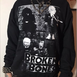 May include: Black sweatshirt with a white graphic print of a band called "BROKEN BONES". The graphic features a black and white photo of a band with a brick wall in the background. The person wearing the sweatshirt has a silver chain necklace with a key and handcuffs pendant.
