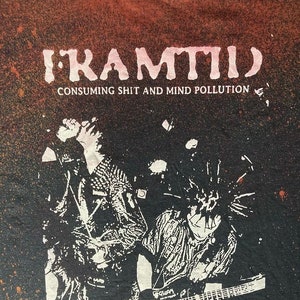 Peut inclure: T-shirt noir avec un design vieilli. Le texte "FRAMTID" est au-dessus de "CONSUMING SHIT AND MIND POLLUTION". Le graphisme présente deux personnages avec des guitares en noir et blanc.