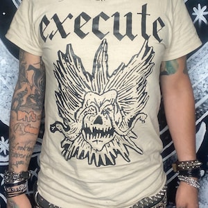May include: Beige t-shirt with the word "EXECUTE" in bold black letters above a detailed black graphic of a skull with wings. The person is wearing a chain necklace and multiple bracelets.