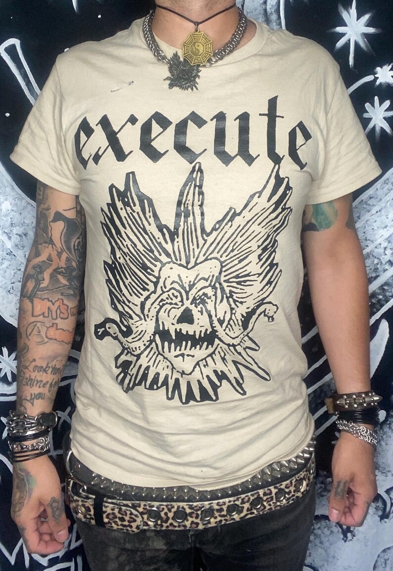 May include: Beige t-shirt with the word "EXECUTE" in bold black letters above a detailed black graphic of a skull with wings. The person is wearing a chain necklace and multiple bracelets.