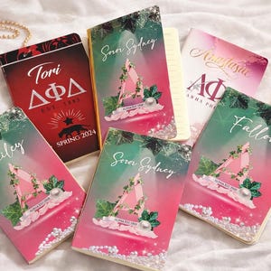 Könnte beinhalten: Fünf rechteckige Notizbücher mit personalisierten Einbänden, die Namen wie Tori, Bailey, Soror Sydney, Anastasia und Fallon tragen. Die Designs zeigen griechische Buchstaben, Efeu, Perlen sowie rosa-grüne Farbverläufe, wobei ein roter Einband den Text DΦΔ aufweist.