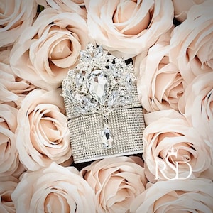 May include: A silver and crystal ring box with a large crystal in the center, surrounded by smaller crystals. The box is sitting on a bed of blush pink roses.