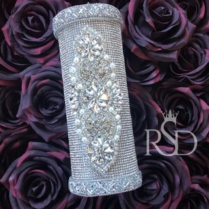 May include: A silver cylinder covered in rhinestones and small white pearls. The cylinder has a decorative design of flowers and leaves. The cylinder is sitting on a bed of dark purple roses.