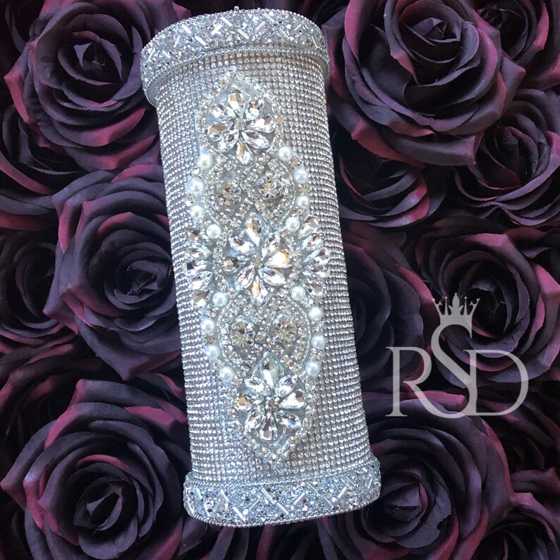 May include: A silver cylinder covered in rhinestones and small white pearls. The cylinder has a decorative design of flowers and leaves. The cylinder is sitting on a bed of dark purple roses.